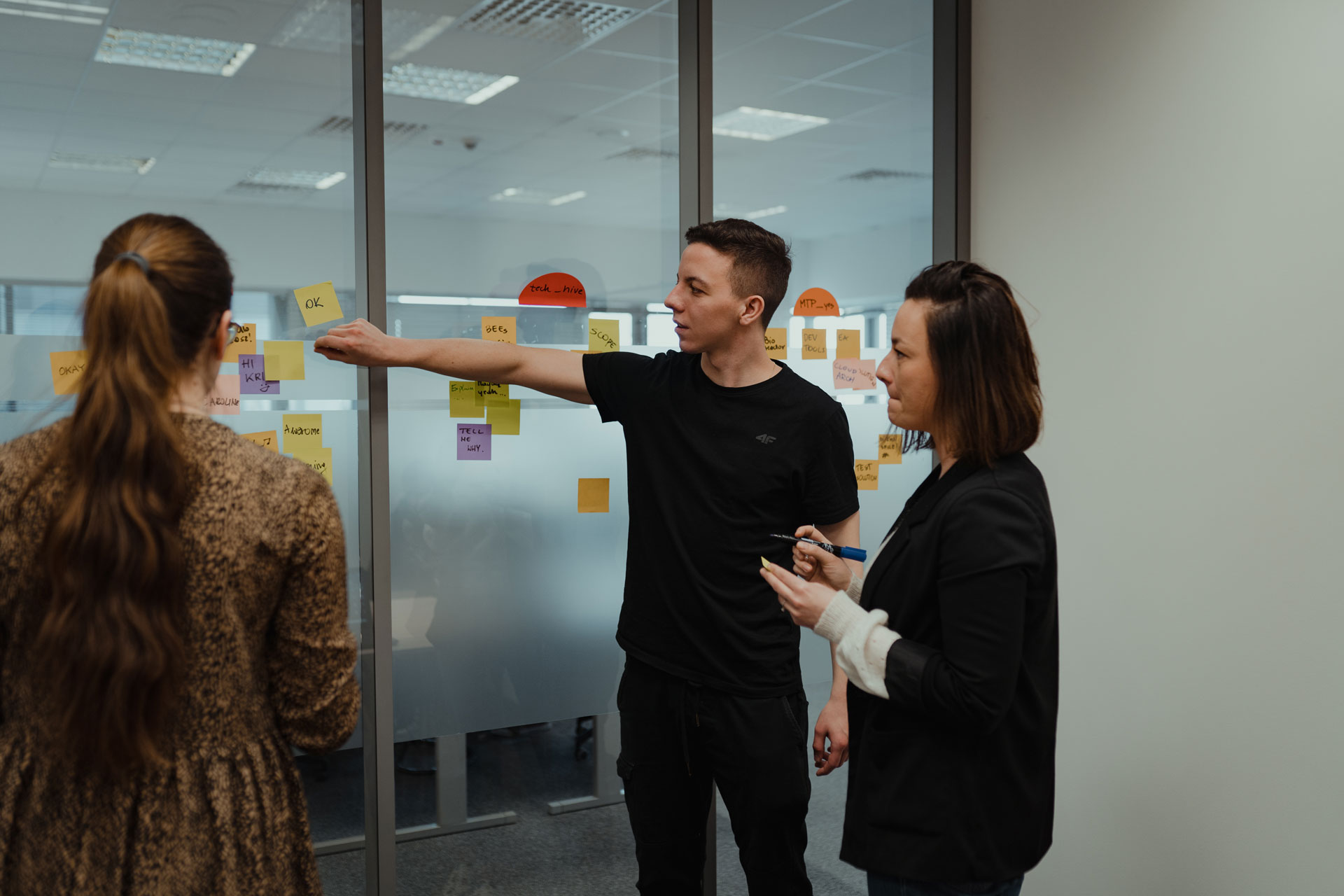 Three people engaged in a discussion over something on a board with post-its