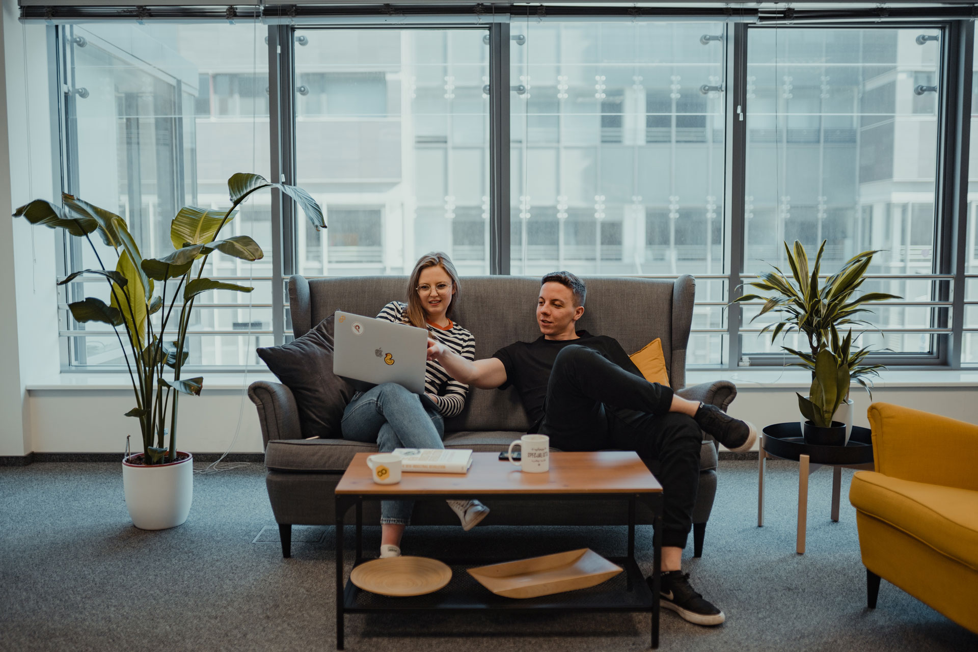 Two people sitting on a couch with a window behind, working together on a laptop