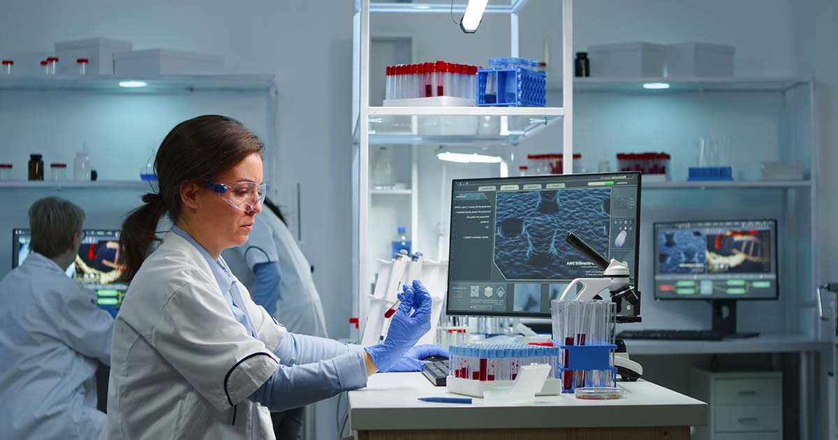 A woman in a lab, looking carefully at a vial with a red liquid in her hand. Two men are looking at a computer screen in the background