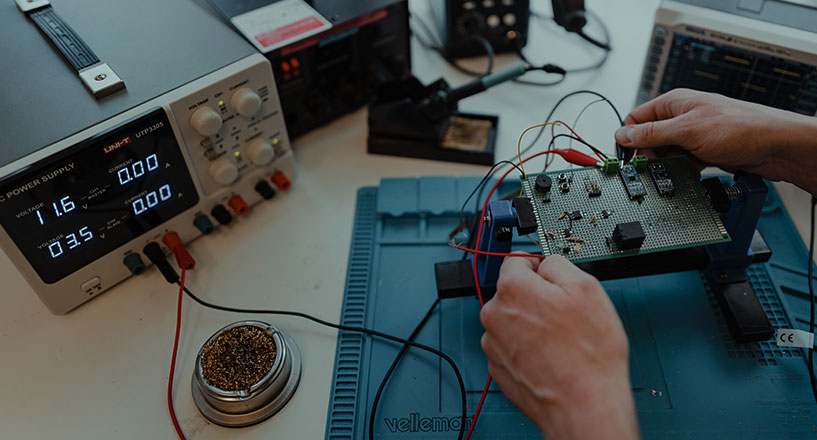 A prototype board connected to a power supply. Two hands reaching from the side, tweaking some components